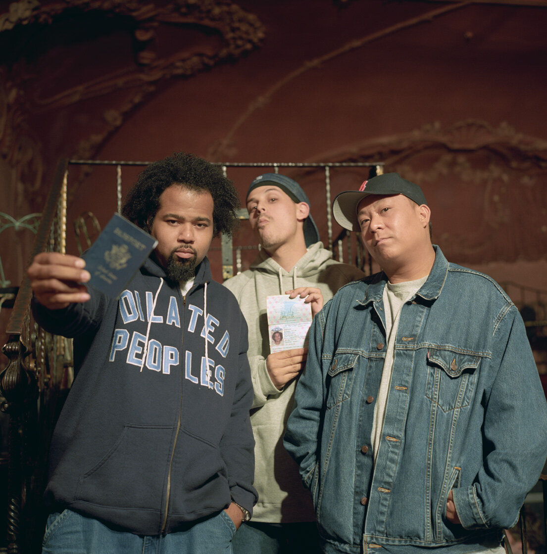 Three men in front of an ornate red wall, one in a Dilated Peoples hoodie holding up a passport