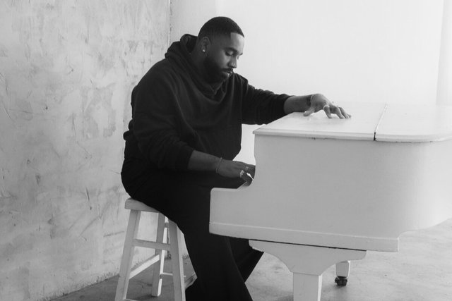 Black and white photo of Dixson seated at a white grand piano in a bare, light-filled room
