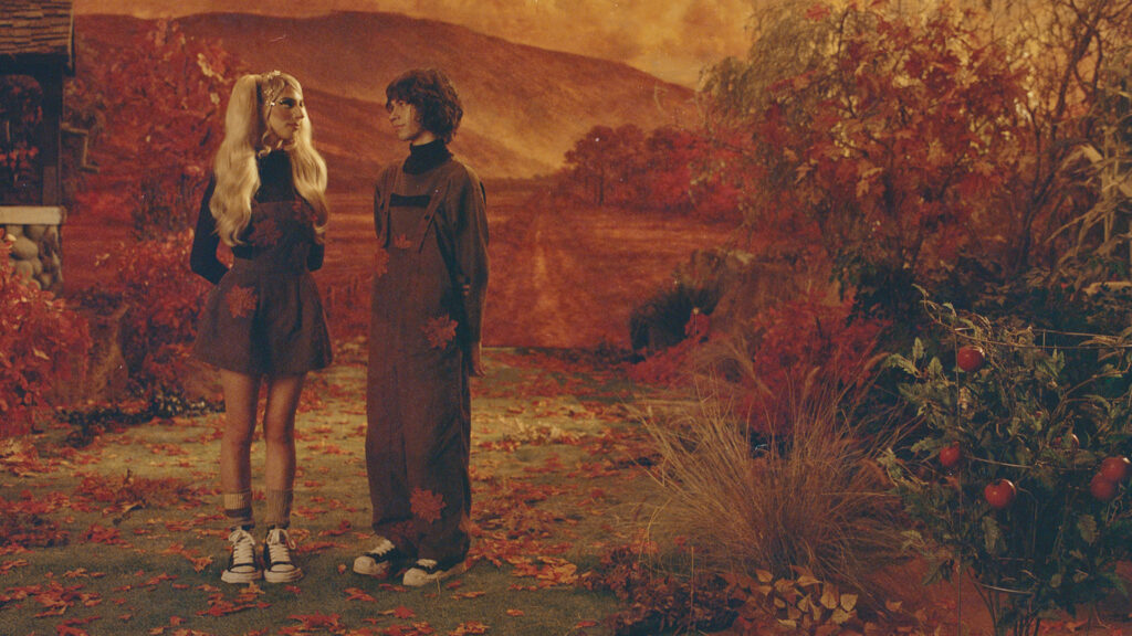 DOMi and JD Beck standing in overalls in an autumn garden of red leaves and tomato plants, the promo image for the Who Asked? Tour