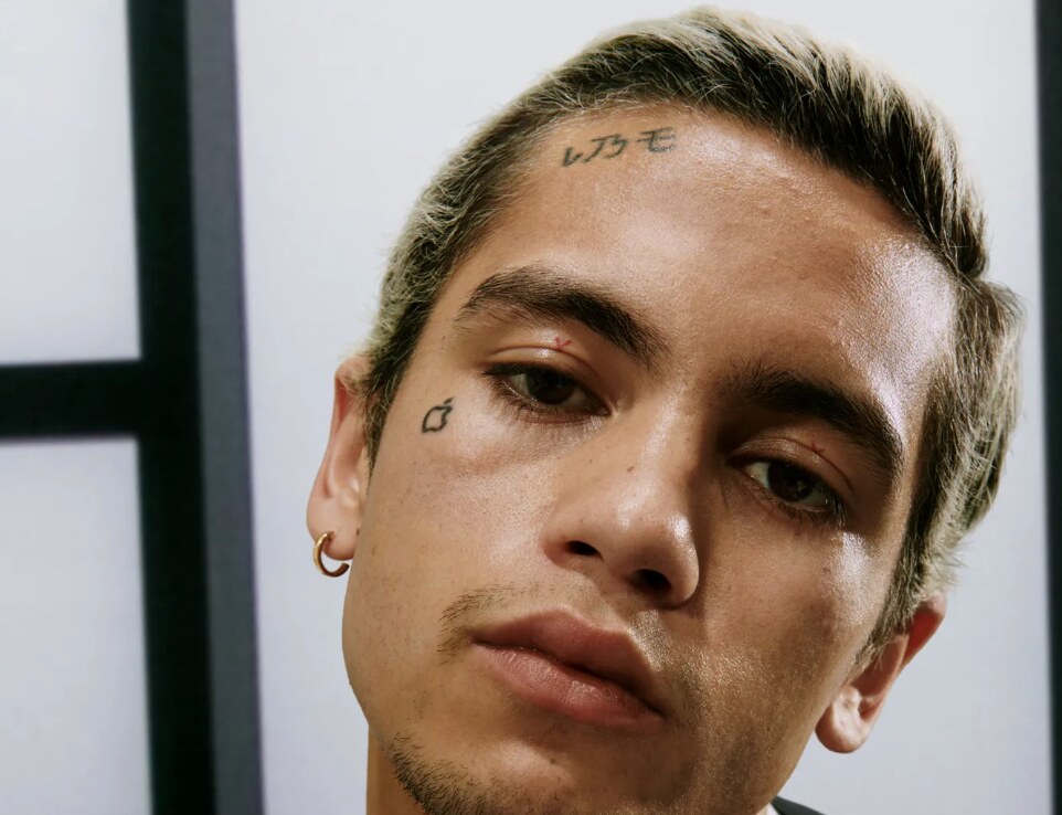 Close-up portrait of Dominic Fike with bleached hair, face tattoos and a gold hoop earring