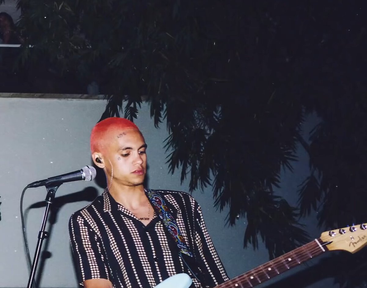 Dominic Fike with bright pink hair playing a Fender electric guitar at a microphone during an outdoor night set