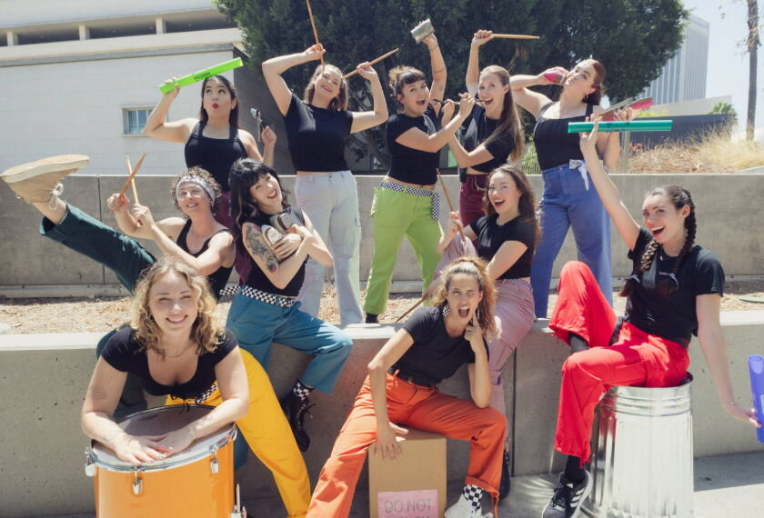Group of smiling performers posing with drumsticks, a snare drum and a trash can drum outdoors