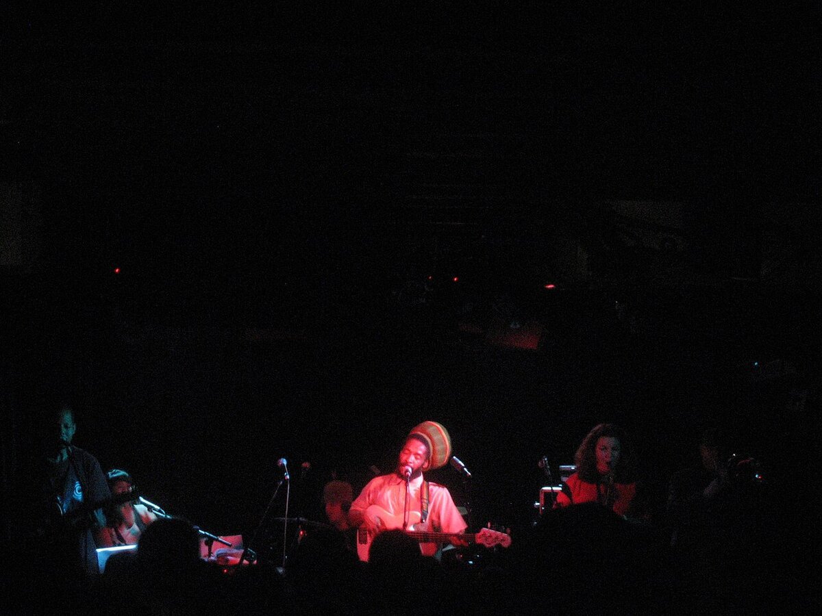 Reggae band on a dark stage lit in red, a bassist in a striped tam singing at the center microphone