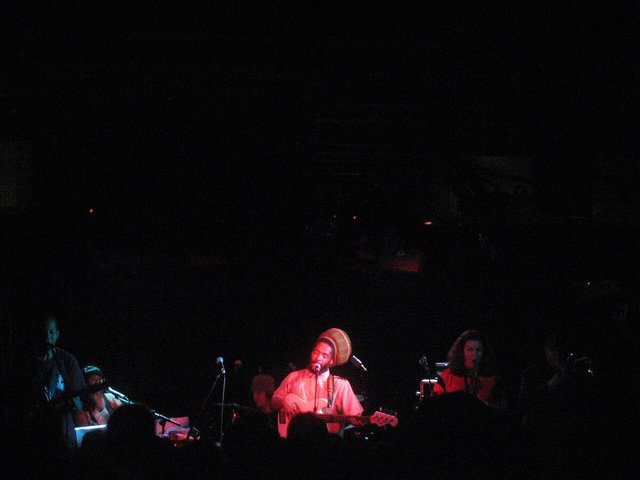 Reggae band on a dark stage lit in red, a bassist in a striped tam singing at the center microphone