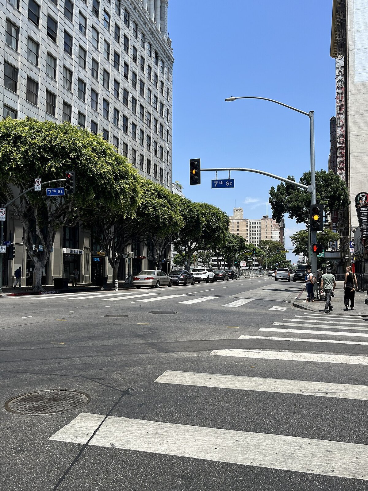 Tree-lined South Main Street at 7th Street in Downtown Los Angeles on a sunny day, with a few pedestrians on the corner