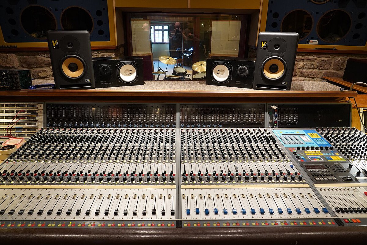 A large recording studio mixing desk with rows of faders and a pair of speaker monitors above it