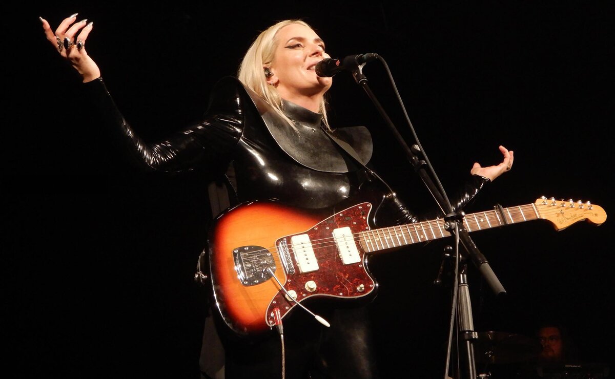 Eivør singing into a microphone on a dark stage with an electric guitar slung across her body