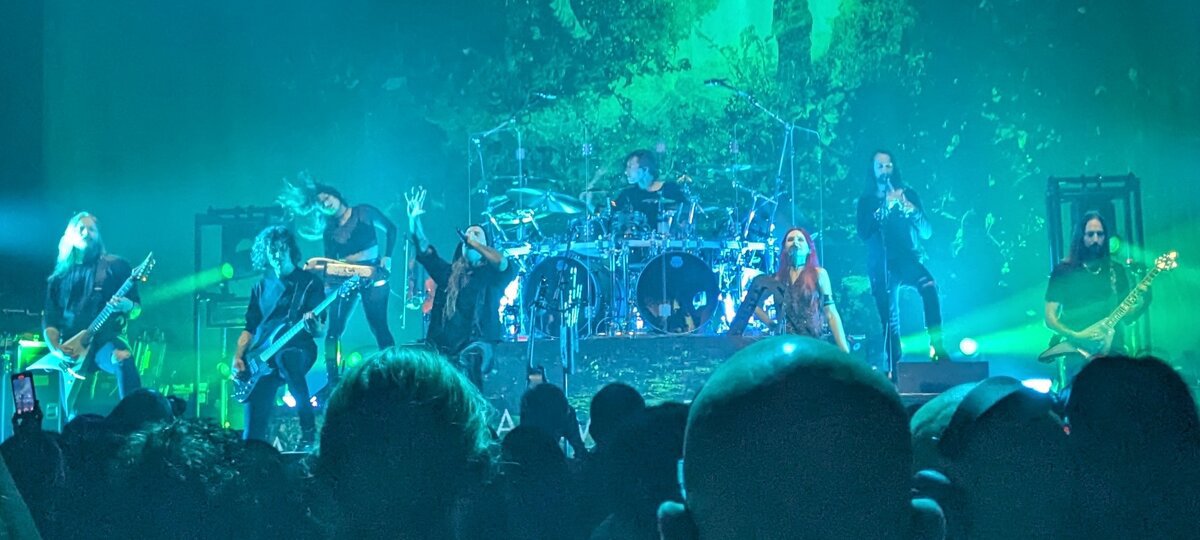 Metal band on a stage lit in green, with guitarists, a drummer and two vocalists, silhouetted crowd heads in the foreground