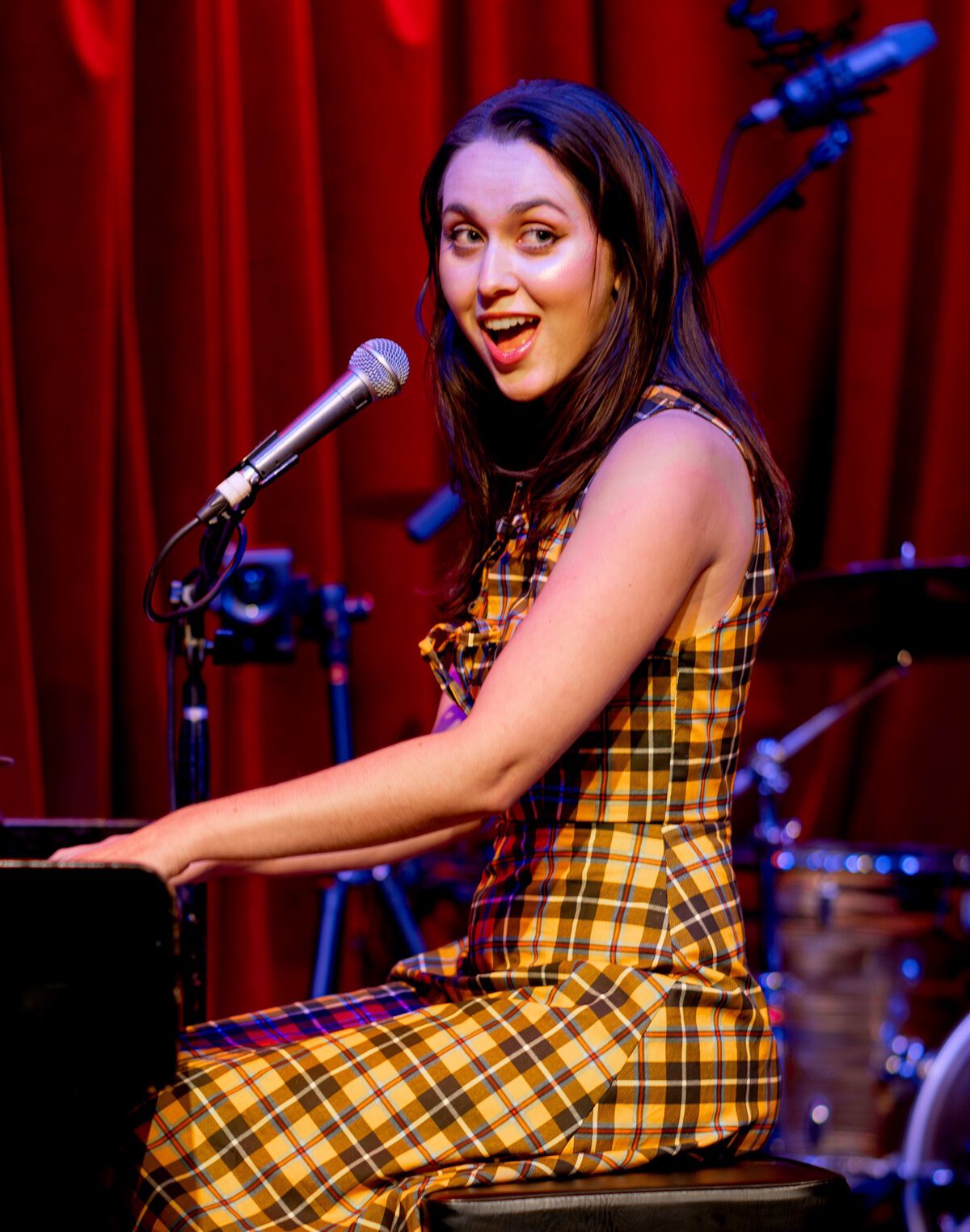 Em Beihold smiling over her shoulder at a microphone while playing piano in front of a red stage curtain