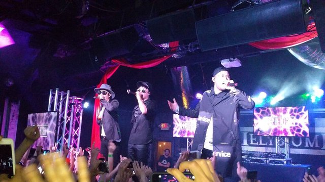 Three members of Epik High performing on a dark club stage in front of a crowd with raised phones
