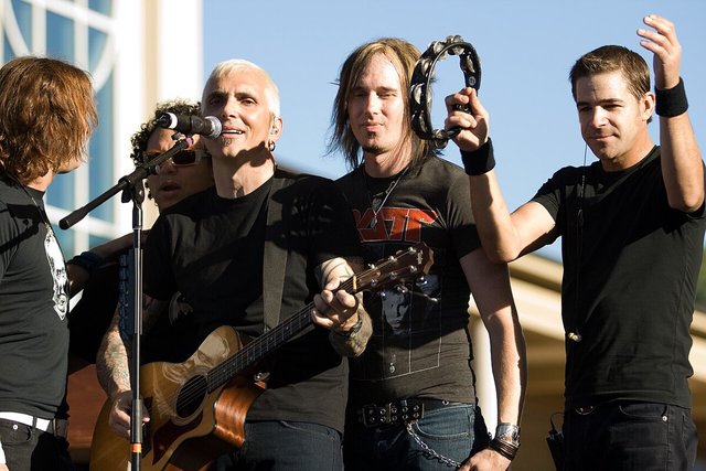 Everclear's lead singer at a microphone with an acoustic guitar, bandmates beside him on an outdoor stage