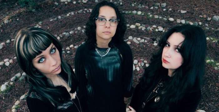 Three members of a band in dark clothing standing among rings of stones, looking up at the camera