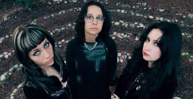 Three members of a band in dark clothing standing among rings of stones, looking up at the camera