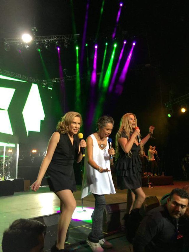 Three women singing into microphones on a stage lit by green and purple beams
