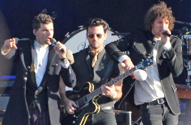 Three members of for KING & COUNTRY singing and playing bass on an outdoor stage in front of a drum kit
