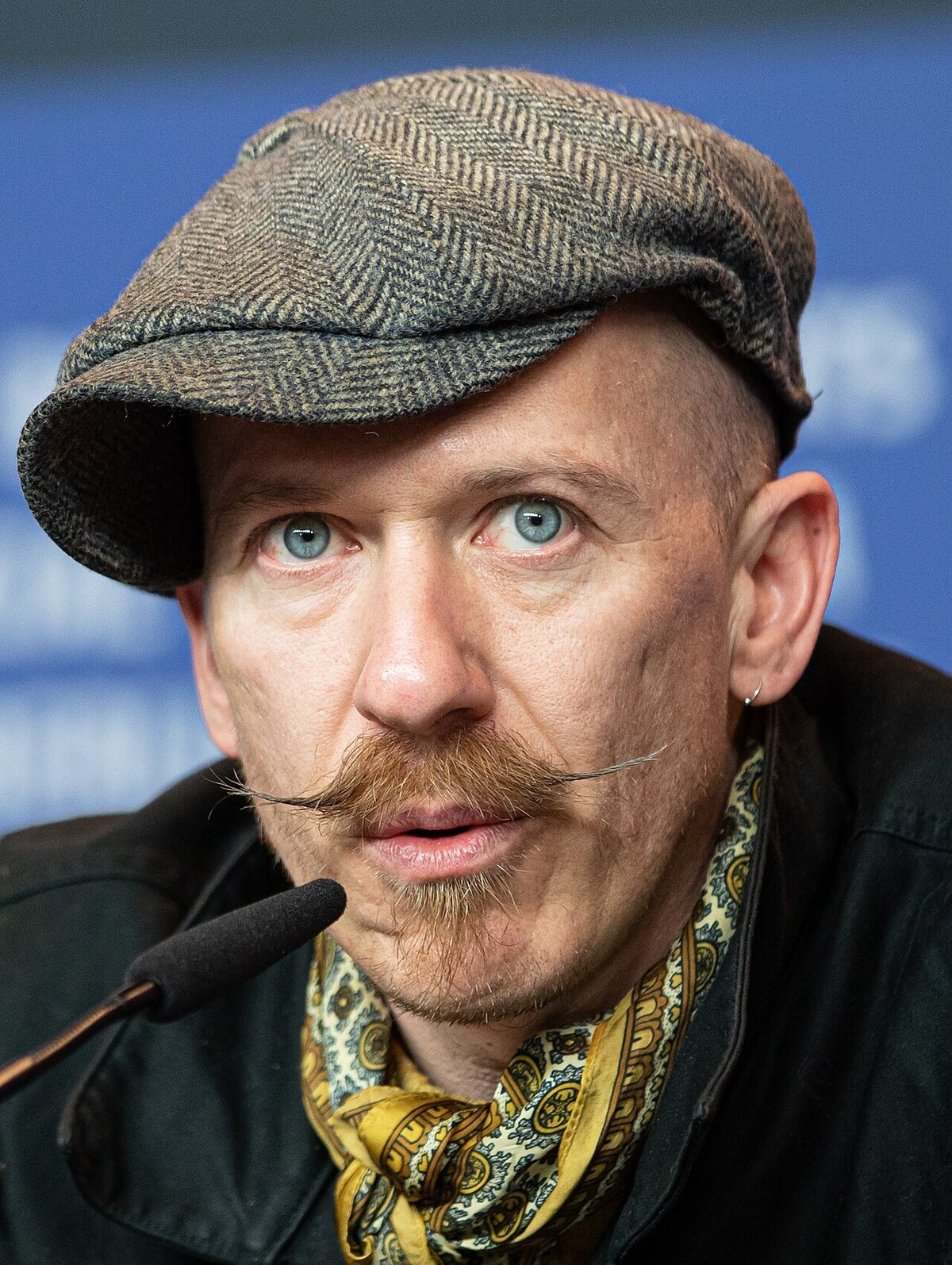 Close-up portrait of a man in a tweed flat cap with a waxed mustache speaking into a microphone
