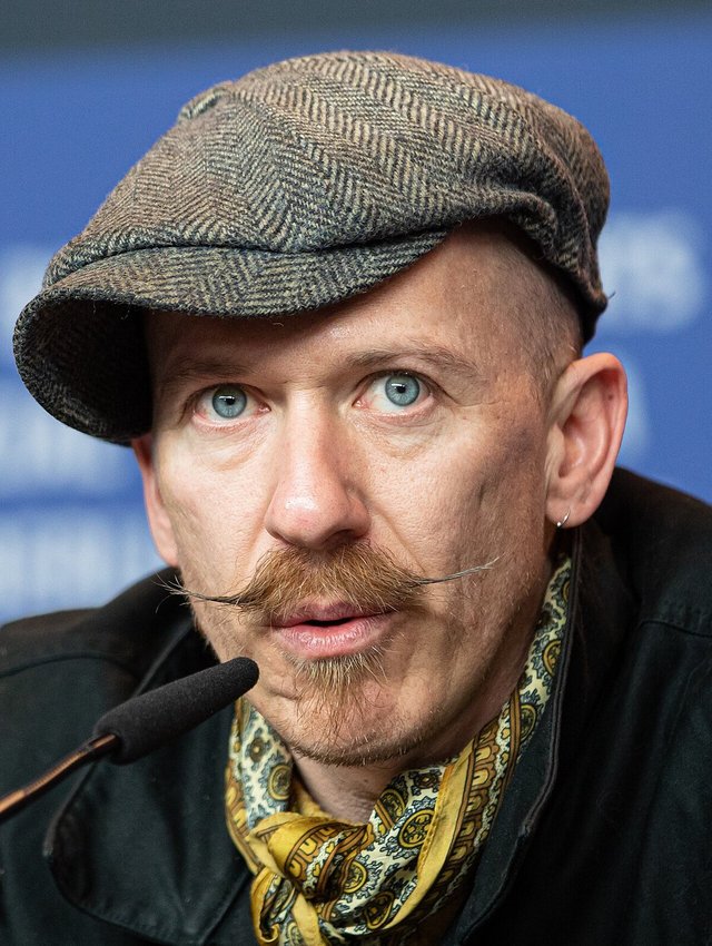 Close-up portrait of a man in a tweed flat cap with a waxed mustache speaking into a microphone