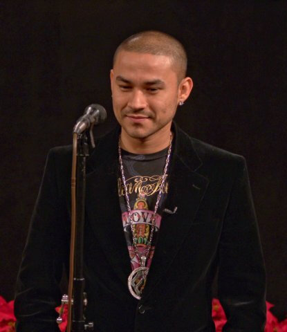 Frankie J in a black velvet jacket and layered chains, smiling slightly beside a microphone stand