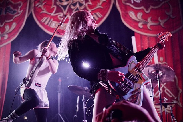 Two guitarists performing on a stage framed by red patterned curtains, one in the foreground playing a bass under purple light