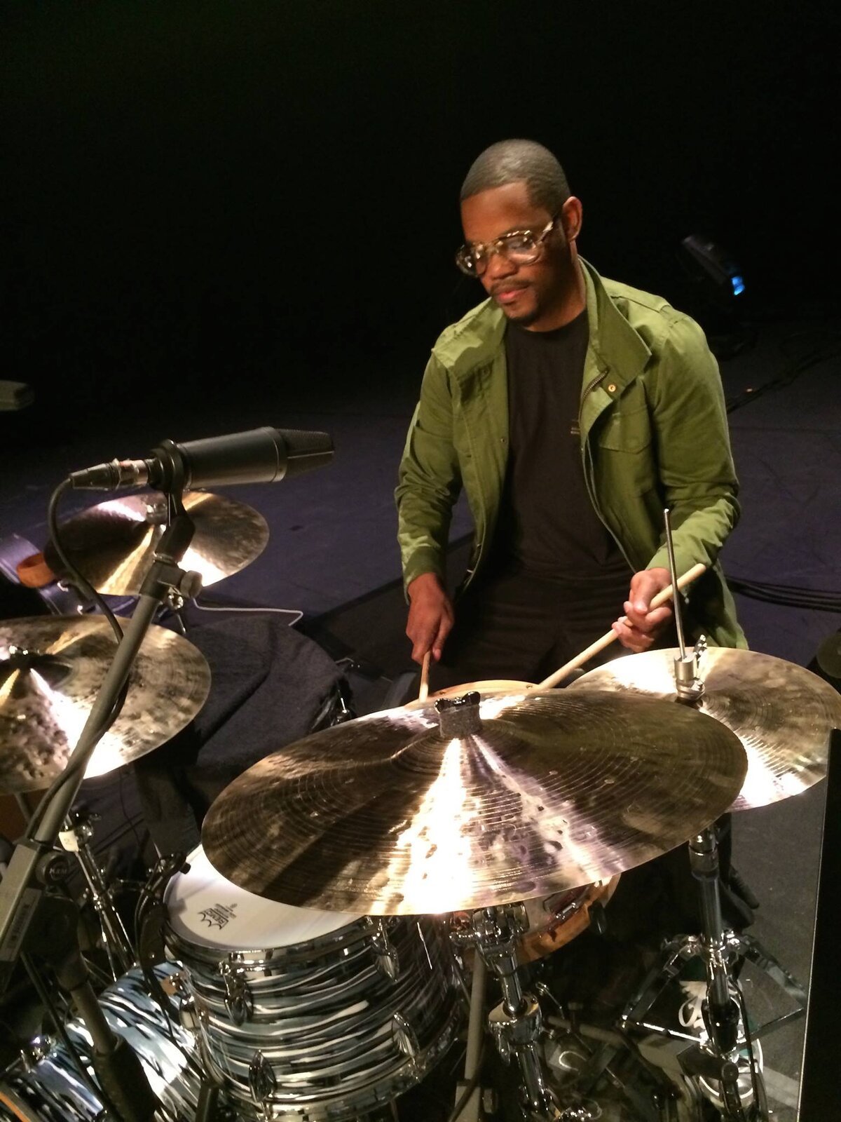 Drummer Karriem Riggins in a green jacket and glasses seated behind a cymbal and drum kit on a dark stage