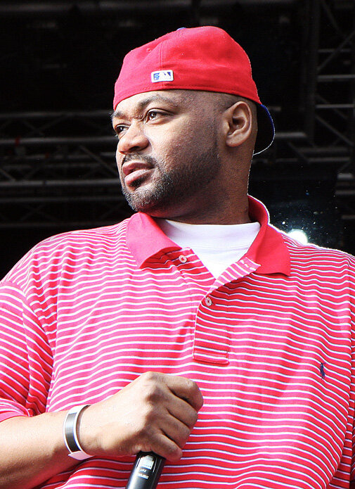 Ghostface Killah holding a microphone on an outdoor stage, wearing a red cap and a red striped polo
