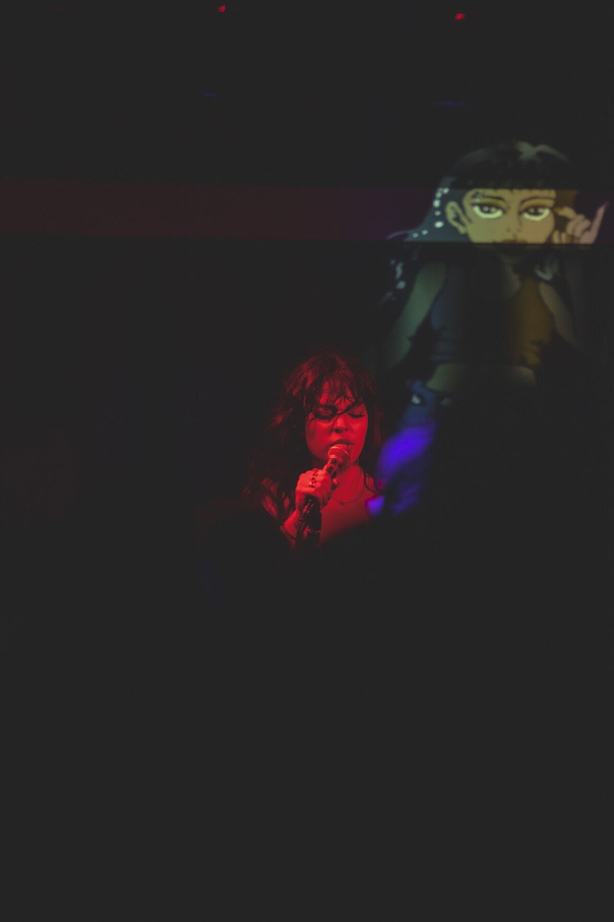 Singer lit in red holding a microphone on a dark stage, with an animated figure projected behind