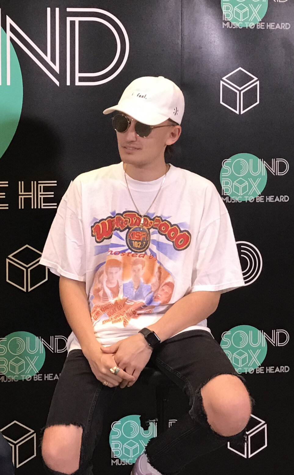 Portrait of gnash seated in a white cap and sunglasses in front of a black step-and-repeat backdrop