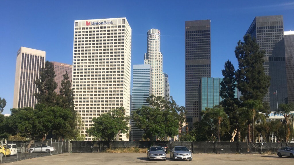 Downtown Los Angeles office towers rising above trees and a parking lot under a clear blue sky
