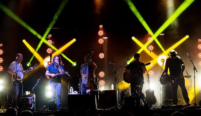Five members of Greensky Bluegrass on a dark stage under crossing green and yellow beams, with banjo, mandolin, upright bass and guitars