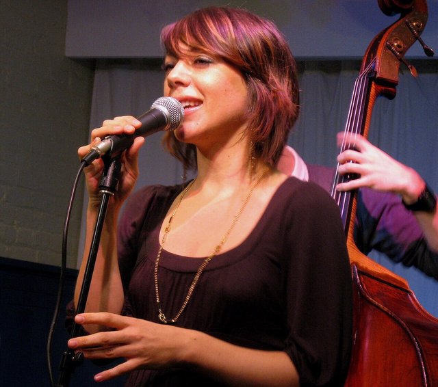 Gretchen Parlato singing into a microphone on stage, with a double bass behind her