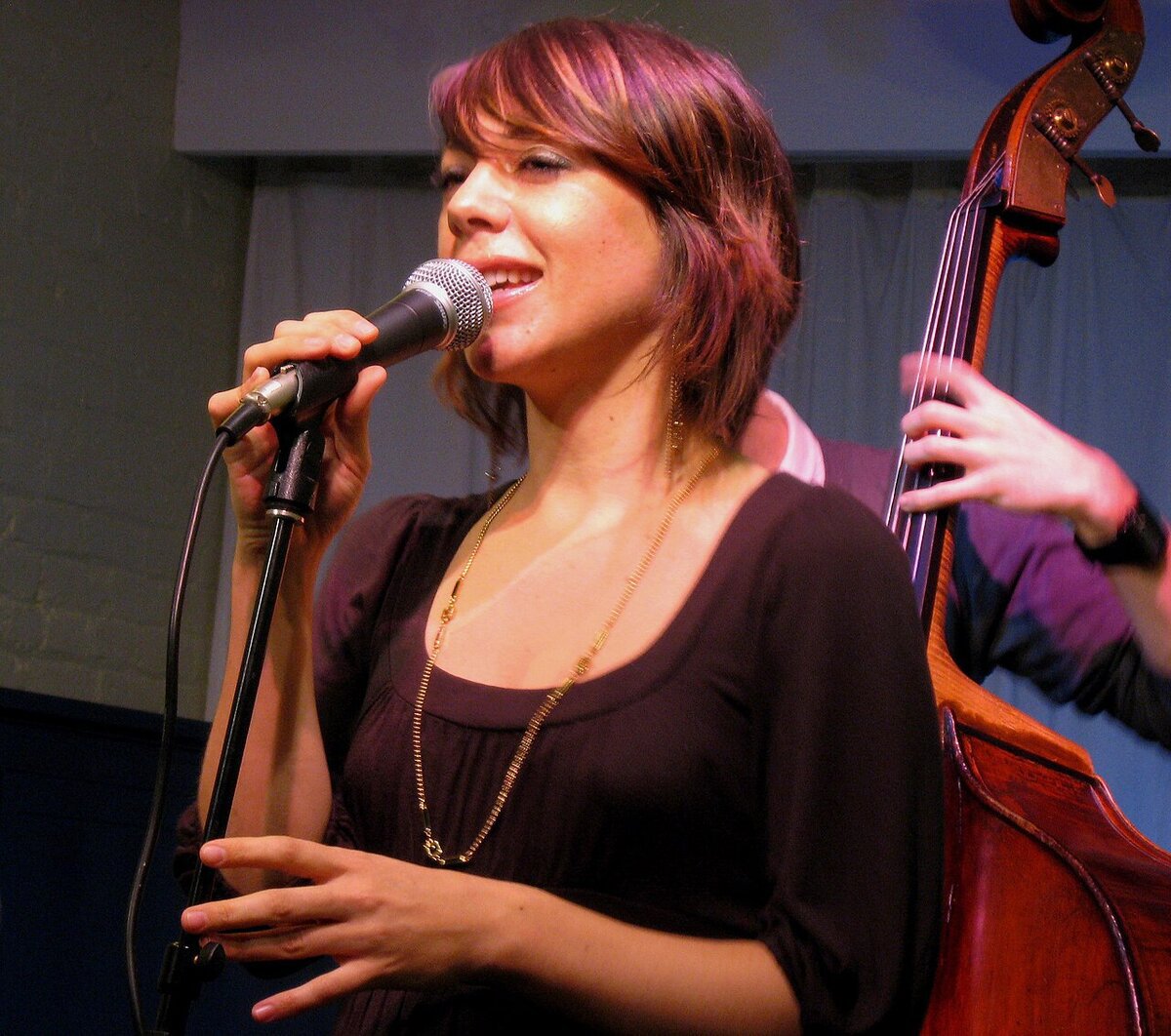 Gretchen Parlato singing into a microphone on stage, with a double bass behind her