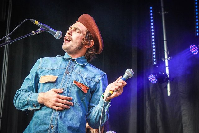 Singer in a denim shirt and beret at a microphone on a dark stage lit with blue lights