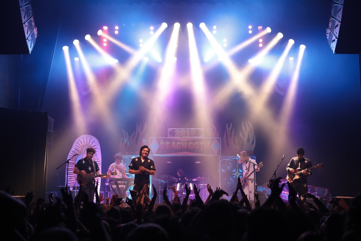 Band playing on a lit stage under beams of purple and white light, with a crowd's raised hands in the foreground