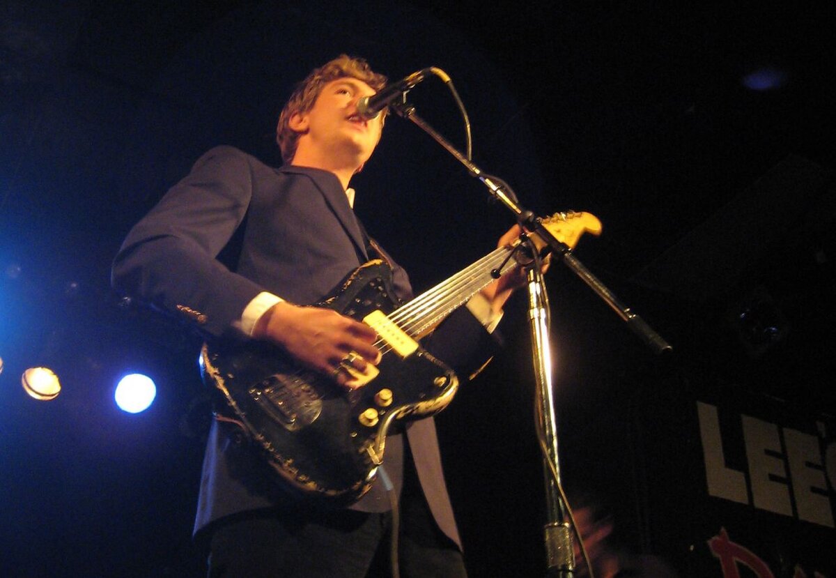 Hamilton Leithauser singing into a microphone while playing a black electric guitar under blue stage lights