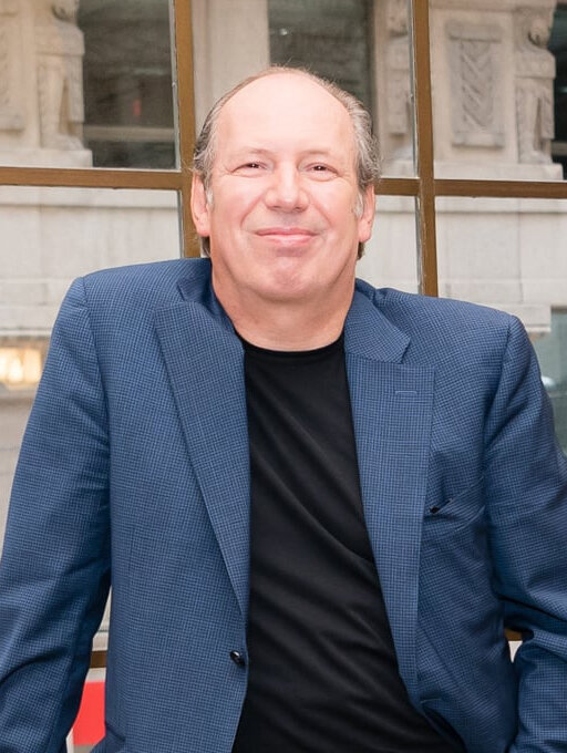 Portrait of Hans Zimmer in a blue blazer and black shirt, smiling in front of a stone building