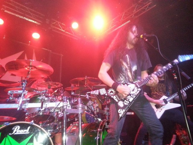 Guitarist in a studded guitar singing into a microphone in front of a drum kit under red stage lights