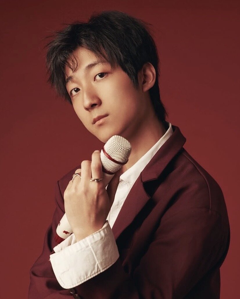 Portrait of HOHYUN in a burgundy blazer holding a white microphone against a red backdrop