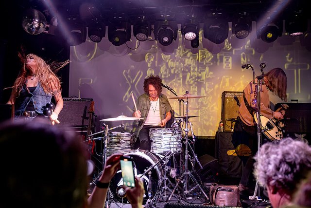 A band with a drummer on a dim club stage under overhead stage lights