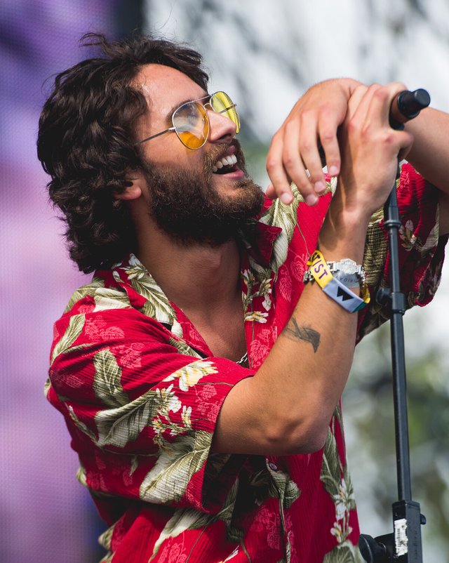 Allan Rayman singing into a microphone on an outdoor festival stage in a red floral shirt and tinted aviator glasses