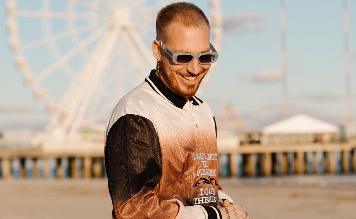 Hugel smiling in sunglasses in front of a Ferris wheel and pier in a promotional portrait