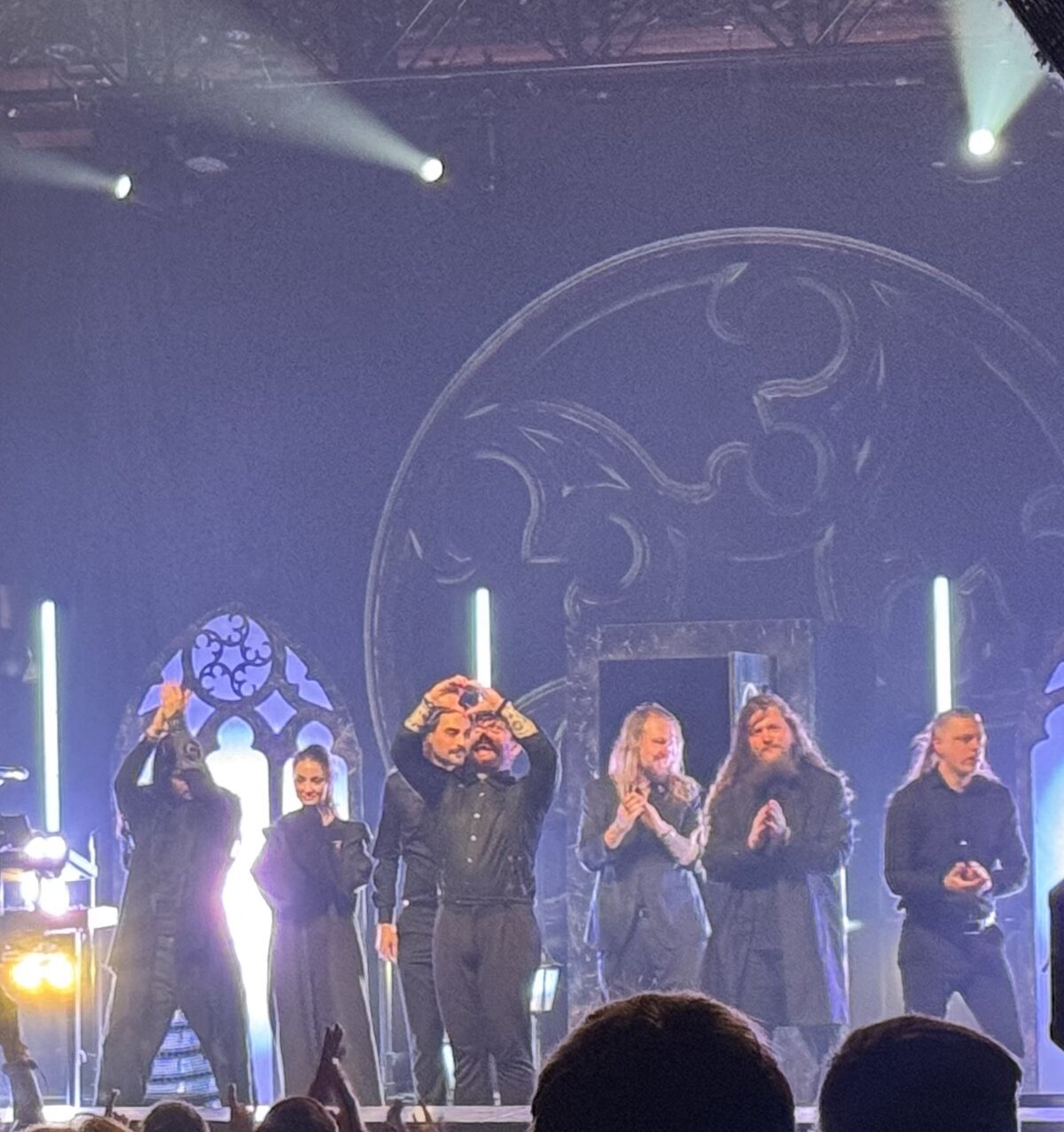 Band members standing on a dimly lit stage with gothic window set pieces, one holding hands in a heart shape