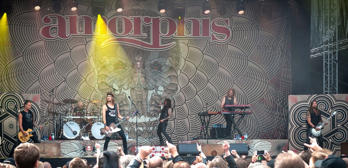 Amorphis performing on a large outdoor stage in front of an ornate backdrop, with a crowd's raised hands in the foreground