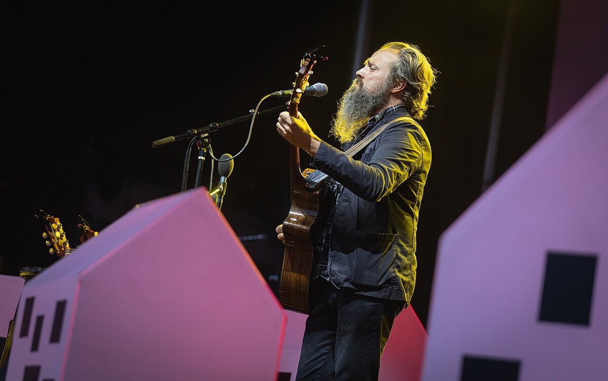 Bearded singer playing an acoustic guitar at a microphone on a dark stage with pink house-shaped set pieces