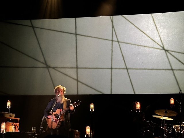 Bearded singer playing an acoustic guitar at a microphone on a dark stage lit by small lamps, with a large geometric projection behind him