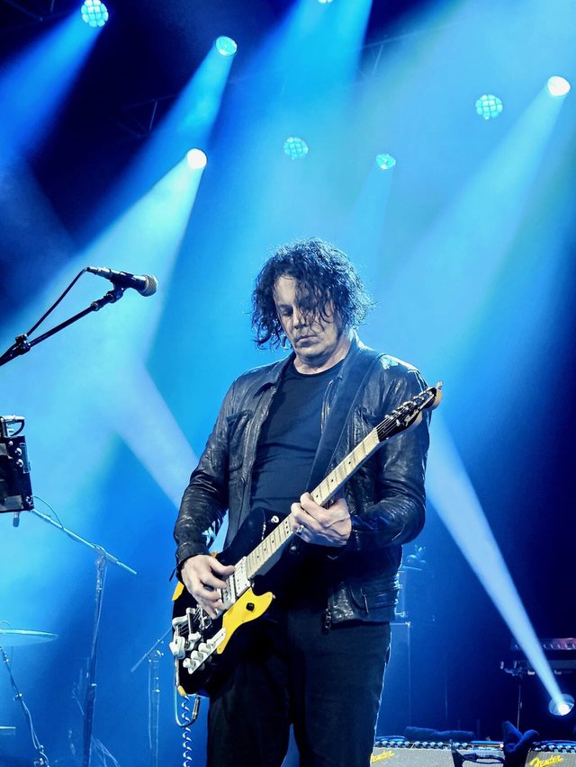 Jack White playing a black and yellow electric guitar under blue stage lights