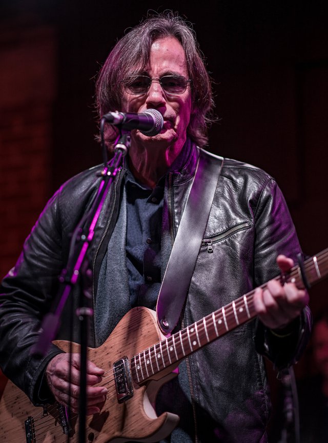 Jackson Browne singing into a microphone while playing an electric guitar, in a black leather jacket under purple stage light