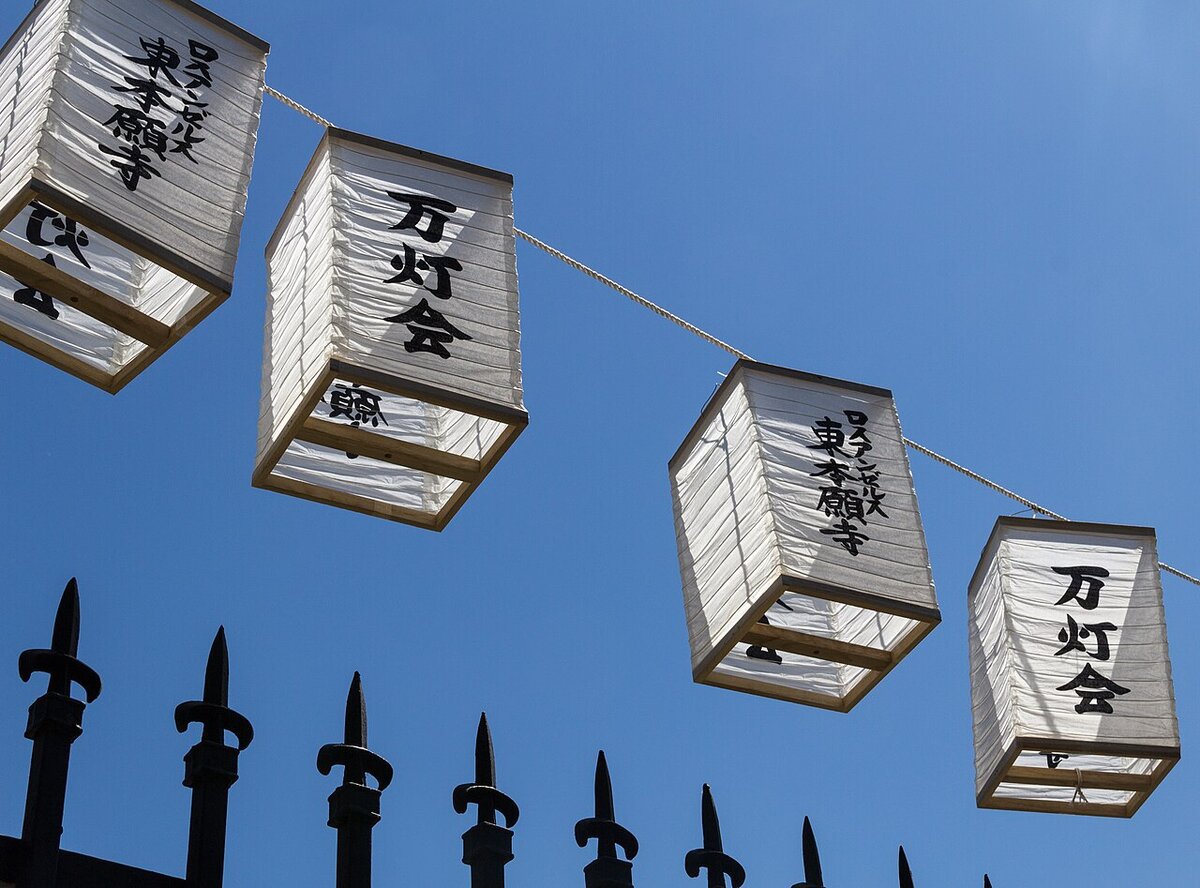 Paper lanterns with black Japanese lettering strung against a blue sky above a spiked iron fence in Little Tokyo