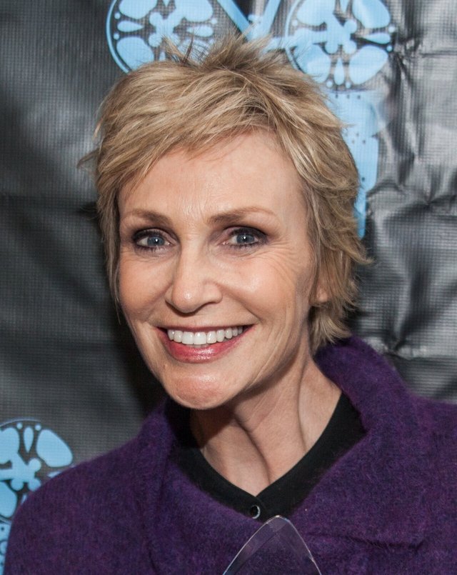 Close portrait of a smiling woman with short blonde hair in a purple jacket in front of a dark backdrop