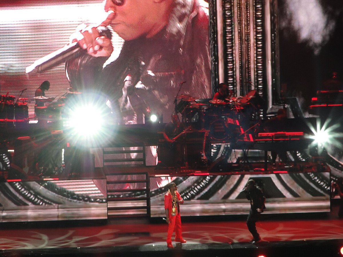 JAY-Z in black rapping into a microphone on a red-lit stadium stage beside Kanye West, with a giant video screen behind them