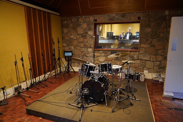 An empty recording studio live room with a drum kit set up in front of a stone wall and a window into the control room